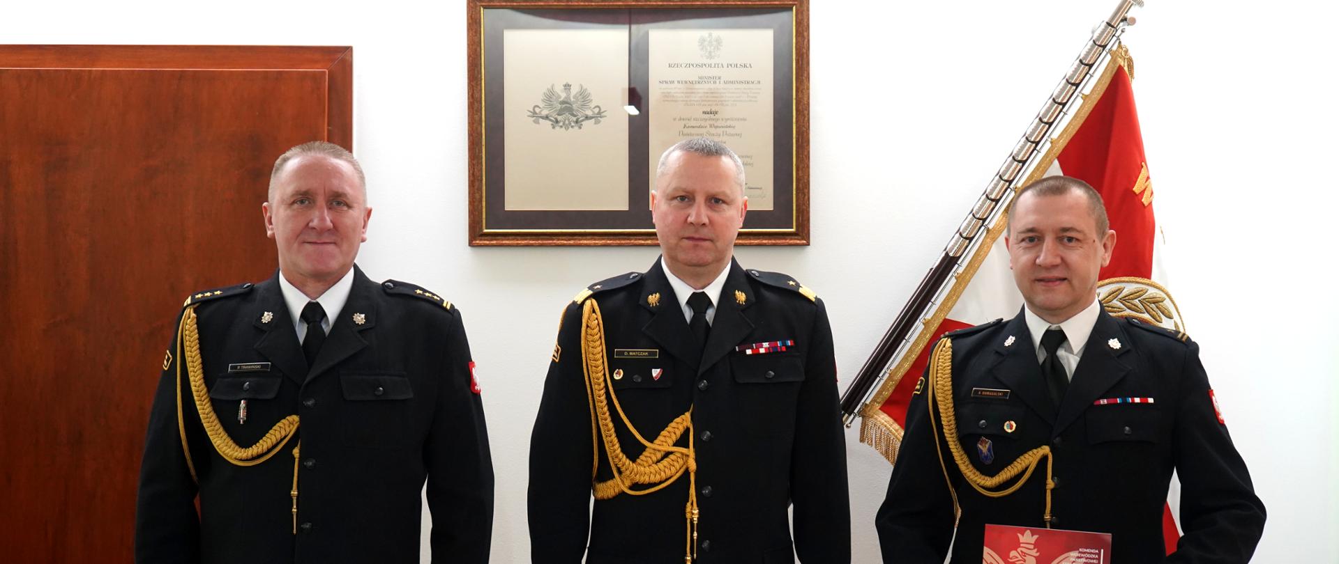 trzech strażaków w ciemnych mundurach ze sznurem stooi obok siebie, w tle sztandar