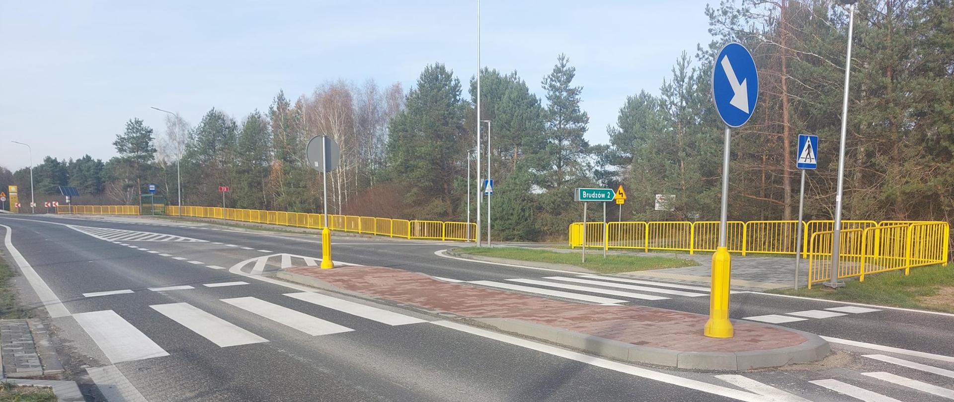 DK73 Lisów - na pierwszym planie przejście dla pieszych z wysepką na środku jezdni. W oddali zatoka autobusowa z wiatą przystankową. od przejścia do przystanku wzdłuż jezdni chodnik oddzielony żółtymi barierkami od przydrożnego rowu 