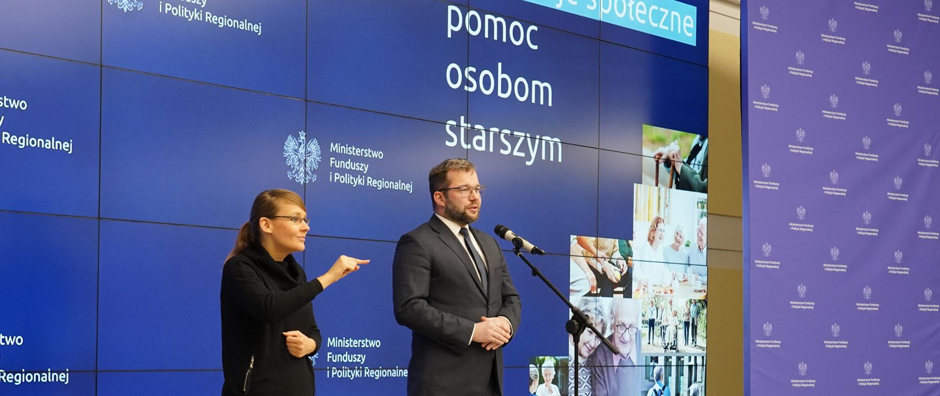 Minister Grzegorz Puda i tłumaczka języka migowego stoją na scenie. Za ich plecami ekran z logo MFiPR oraz napis innowacje społeczne, pomoc osobom starszym.