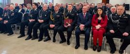 strażacy i druhowie oraz osoby w eleganckich ubraniach siedzą na krzesłach w sali