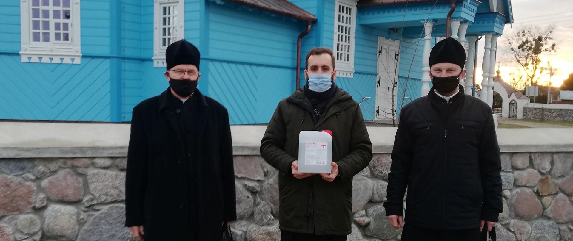 Na tle niebieskiej cerkwi stoi trzech duchownych w sutannach. Jeden z nich stojący po środku trzyma zbiornik z płynem do dezynfekcji.