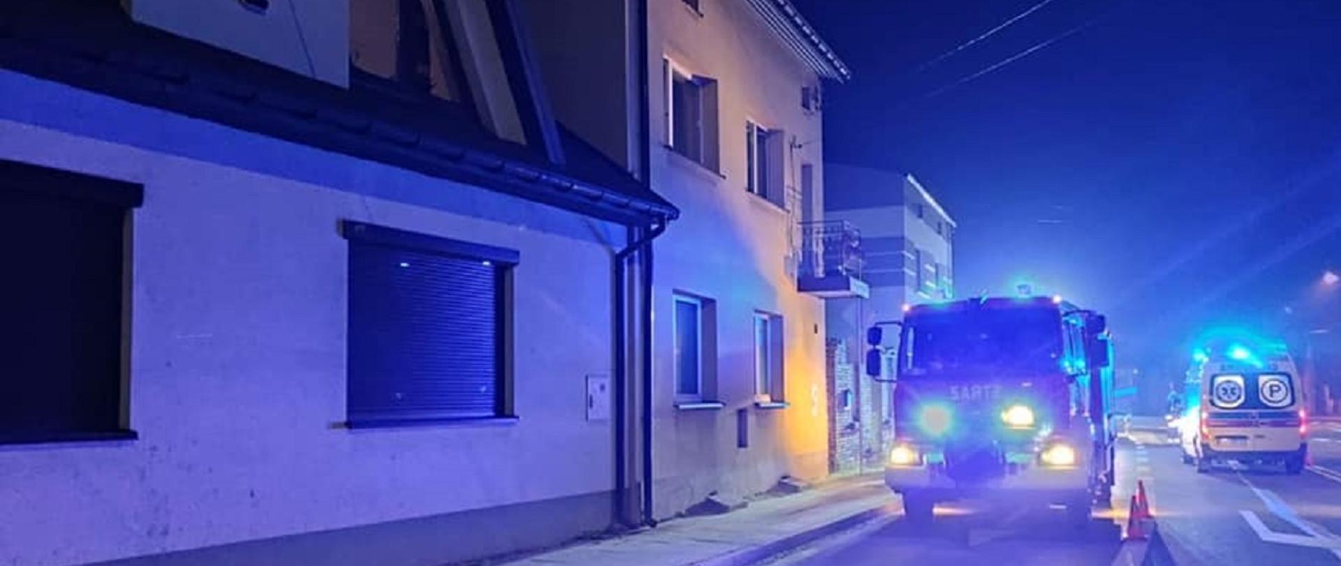 Zdjęcie przedstawia pojazd Straży Pożarnej oraz karetki Pogotowia Ratunkowego stojące na jezdni na sygnałach świetlnych przed budynkiem w którym doszło do wybuchu gazu. Zdjęcie wykonano późnym wieczorem