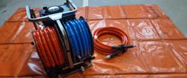 węże hydrauliczne nawinięte na zwijadło koloru czerwonego i niebieskiego obok lezy jeden krąg węza koloru czerwonego wszystko znajduje się na macie koloru pomarańczowego
