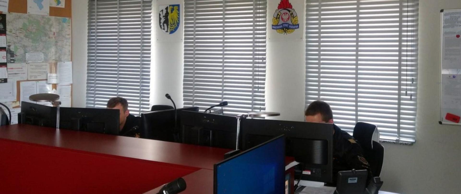Na zdjęciu widoczne jest pomieszczenie Stanowiska Kierowania Komendanta Miejskiego PSP w Bytomiu. Za pulpitem widać monitory komputerów i siedzących dwóch strażaków - dyżurnych w umundurowaniu służbowym. Na ścianach widoczne logo PSP i herb Bytomia.