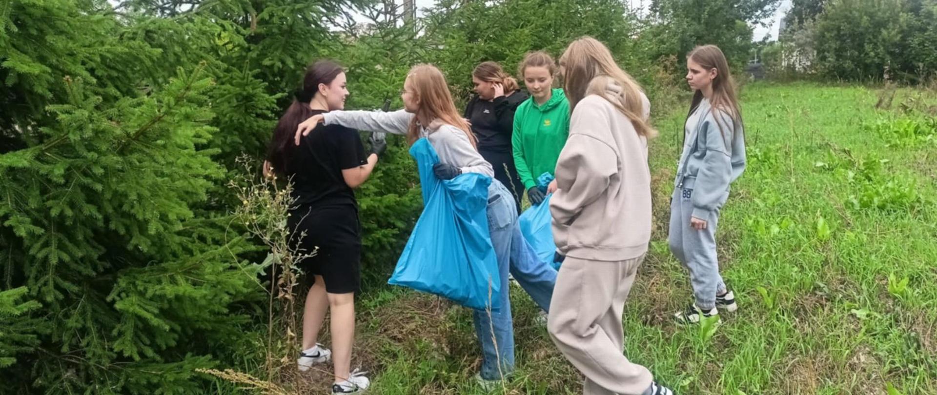 Na zdjęciu widać grupę siedmiu osób na zielonej, trawiastej polanie w pobliżu świerkowych drzewek. Kilka z nich trzyma duże niebieskie worki na śmieci i ma w dłoniach czarne rękawiczki ochronne. Jedna dziewczyna w jasnej bluzie z wyciągniętą ręką pokazuje coś w stronę gałęzi, obok stoi osoba w czarnym stroju, a reszta grupy przygląda się pozostałym osobom.