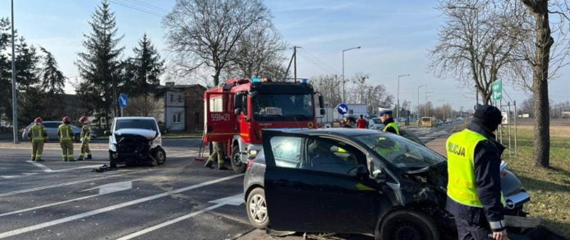 Po prawej widać ciemny uszkodzony samochód, drzwi są otwarte, po lewej biały uszkodzony samochód, uszkodzony przód, za pojazdami samochód strażacki