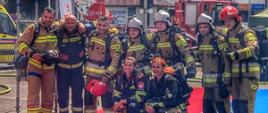 Zdjęcie wykonane podczas zawodów Najtwardszy Strażak. Na fotografii grupowe zdjęcie kilku strażaków podczas zawodów w Rzeszowie. Strażacy ubrani w umundurowania specjalne bojowe. Zdjęcie wykonane podczas zawodów przed budynkiem Urzędu Wojewódzkiego w Rzeszowie na tle tablicy z wynikami po zakończonej konkurencji. Z tyłu widać samochód strażacki typu podnośnik. 