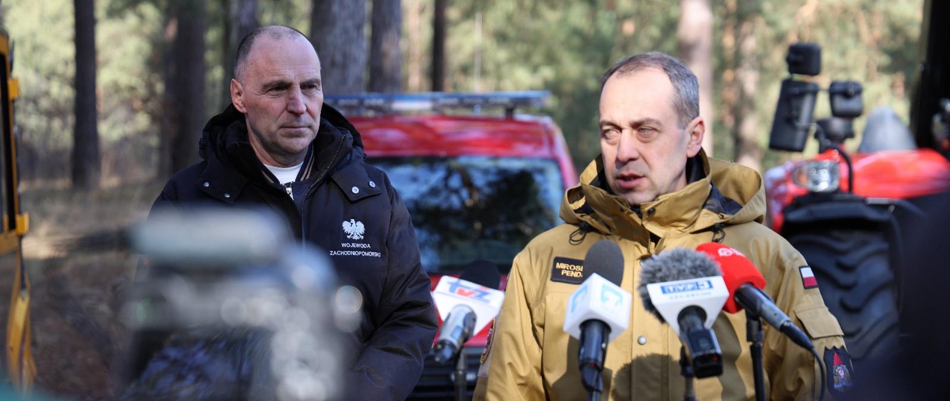 Dwóch mężczyzn stoi w lesie przed mikrofonami i udziela wypowiedzi mediom. Za nimi widać czerwony samochód straży pożarnej.
