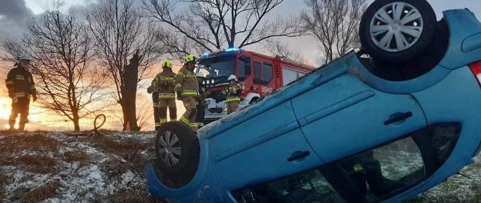 Niebieski samochód leży na dach poza drogą na poboczu w tle widoczny samochód straży pożarnej oraz strażacy w mundurach koloru piaskowego oraz czarnego w oddali widoczne drzewa 