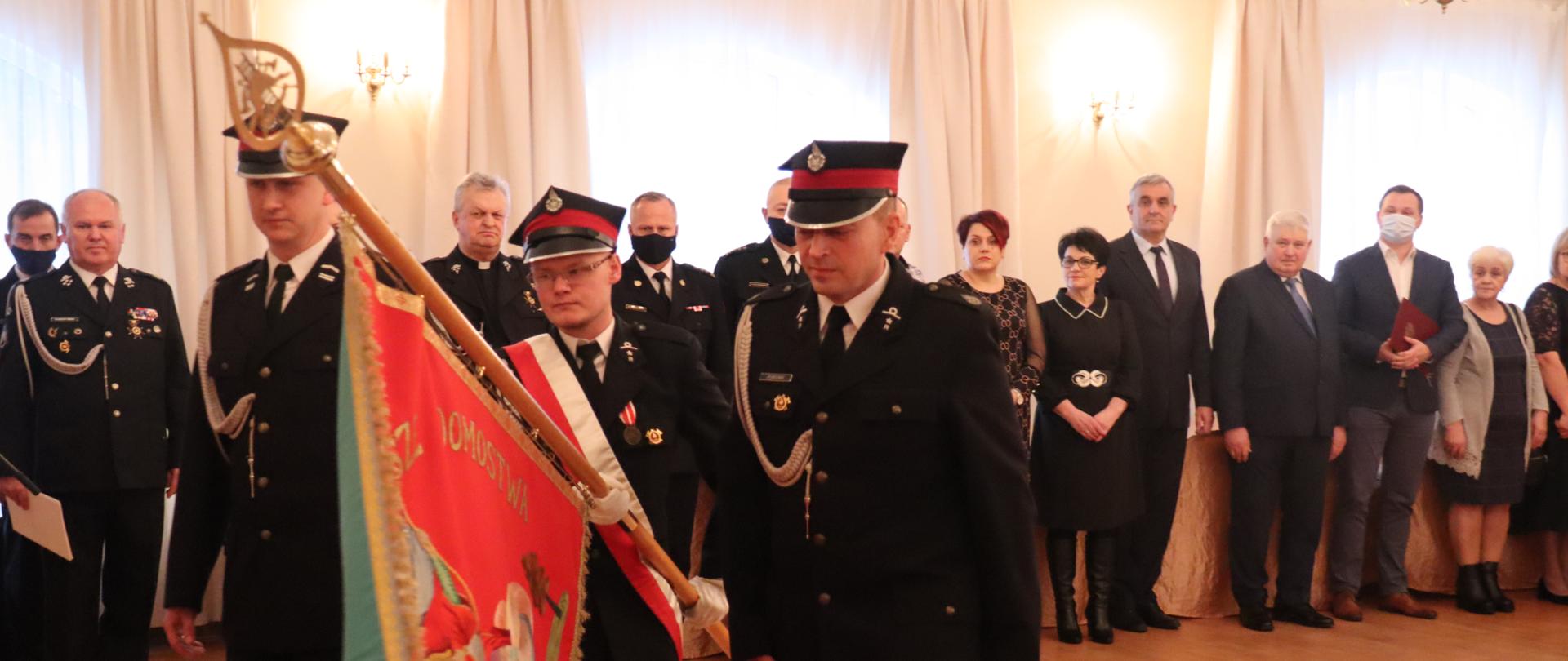 Na pierwszym planie poczet sztandarowy jednostki OSP. W tle szereg gości zaproszonych na uroczystość. Rzecz dzieje się w sali.
