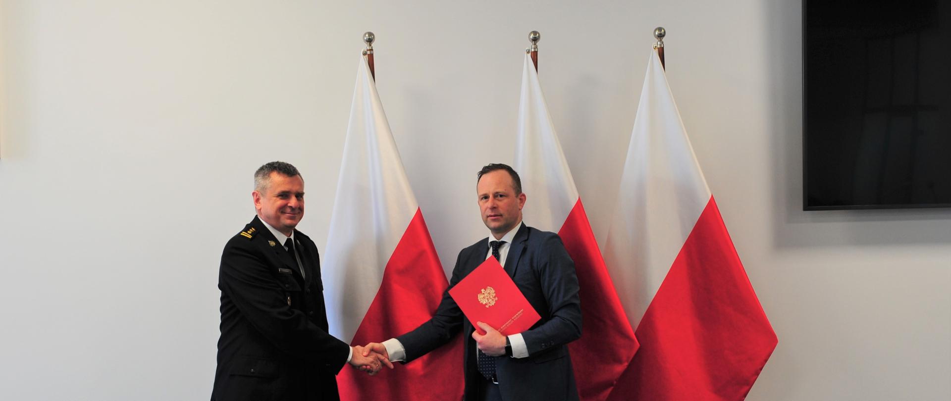 Pomorski komendant wojewódzki PSP uściska dłoń z prezesem Wojewódzkiego Funduszu Ochrony Środowiska i Gospodarki Wodnej w Gdańsku na tle flagi państwowej oraz godła Rzeczypospolitej Polskiej. Prezes Wojewódzkiego Funduszu Ochrony Środowiska i Gospodarki Wodnej w Gdańsku trzyma podpisany dokument. 