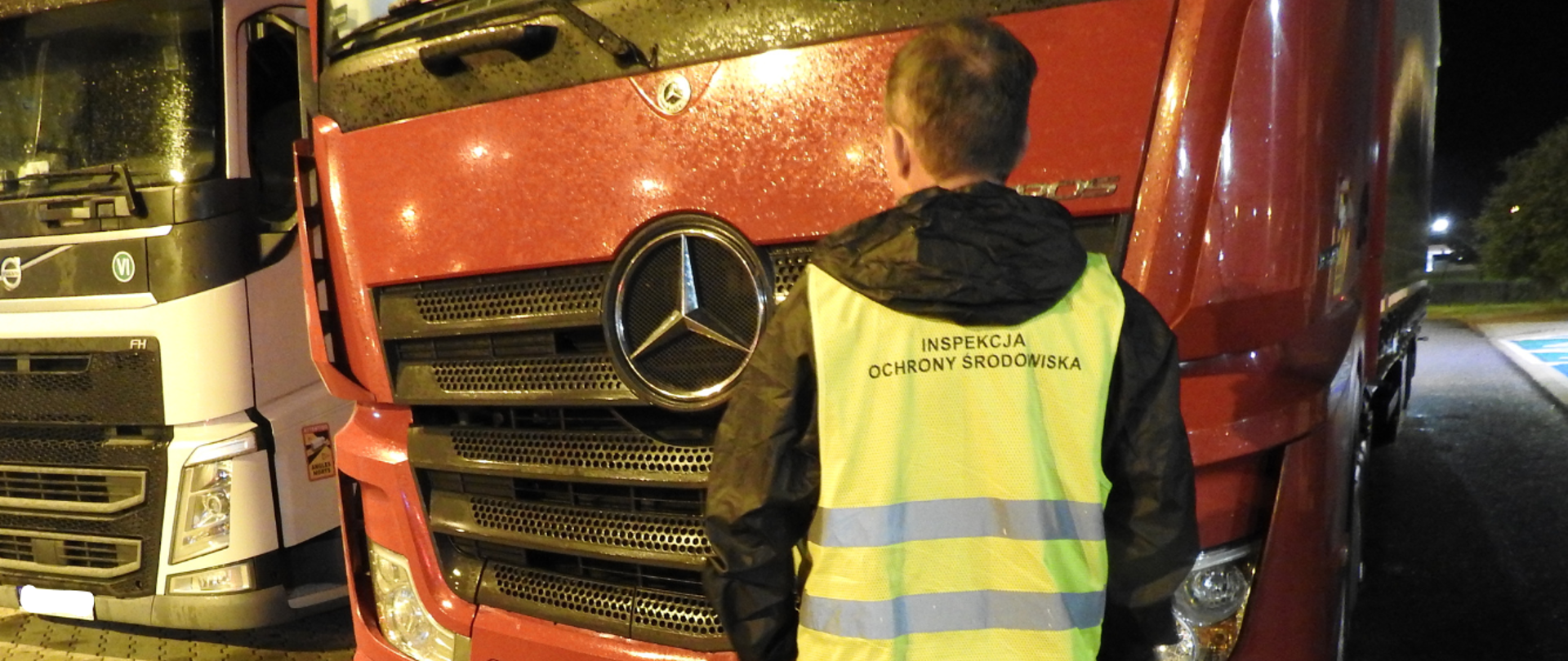 Mężczyzna stojący tyłem do zdjęcia, ubrany w odblaskową kamizelkę z napisem Inspekcja Ochrony Środowiska. Przed mężczyzną stoją zaparkowane samochody ciężarowe.