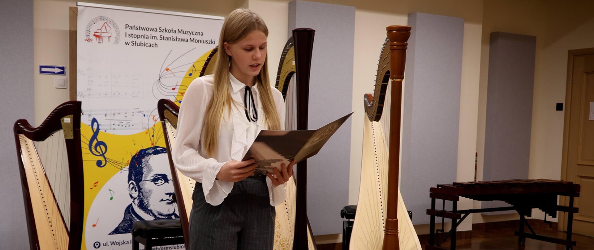Uczennica stoi w auli szkolnej i czyta plan koncertu.