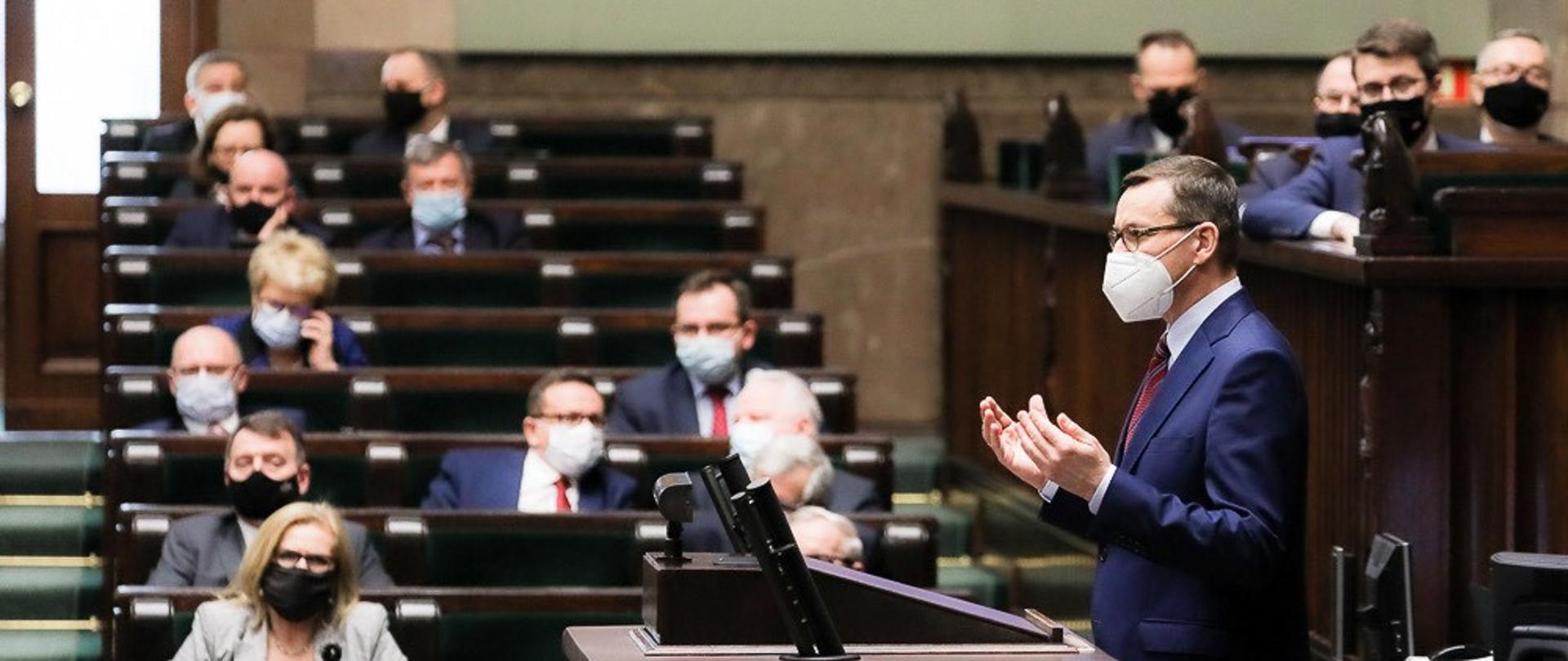 Premier Mateusz Morawiecki stoi przy mównicy w Sejmie. Na drugim planie posłowie siedzący w ławach sejmowych.