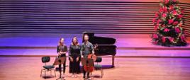 Dwie kobiety i mężczyzna stoją obok siebie na estradzie sali koncertowej , za nimi stoi fortepian. po prawej stronie z tyłu stoi udekorowana choinka.