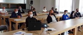 Widok z przodu. Za stołami na sali OSP siedzą uczestnicy spotkania w maseczkach ochronnych. 