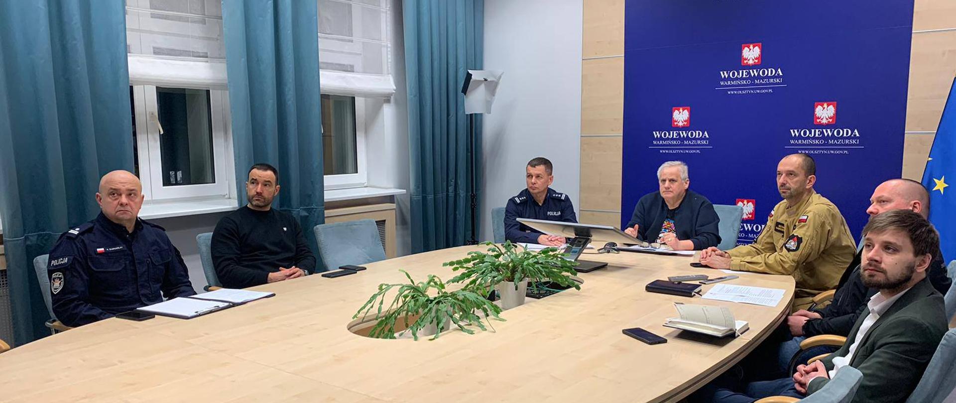 Grupa ludzi siedzi przy stole, mają poważne miny patrzą w jeden punkt - na wideokonferencje