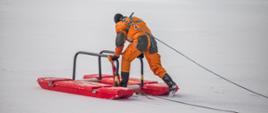 Zdjęcie przedstawia strażaka ubranego w ubranie wypornościowe koloru pomarańczowego, przy pełnej asekuracji, idący z trapem ratowniczym po lodzie na akwenie.
