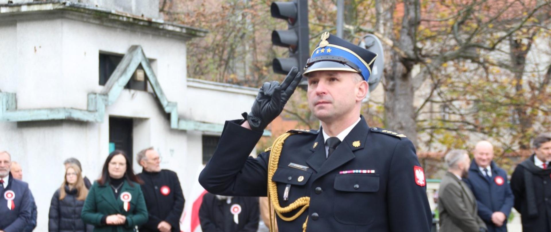 Na zdjęciu Komendant Miejski Państwowej Straży Pożarnej st. bryg. Tomasz Polak w umundurowaniu wyjściowym. Oddaje honor po złożeniu kwiatów pod pomnikiem marszałka Józefa Piłsudskiego. Zdjęcie wykonane w ciągu dnia. Zachmurzenie całkowite.