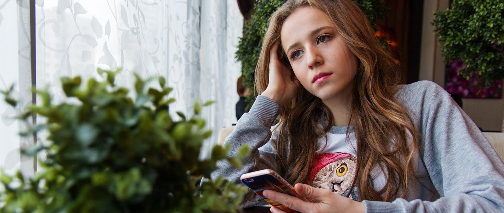 Zamyślona nastolatka siedzi przy stole, w reku trzyma smartfona.