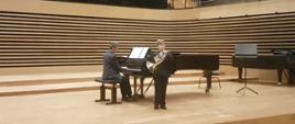Uczeń stoi przed fortepianem i gra na waltorni, nauczyciel gra na fortepianie na scenie sali koncertowej PSM