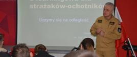 Inauguracja szkolenia podstawowego W świetlicy komendy powiatowej PSP, na tle ekranu, na którym wyświetlany jest napis "Platforma e-learningowa dla strażaków-ochotników. Uczymy się na odległość", stoi zastępca komendanta powiatowego PSP w ubraniu służbowym koloru piaskowego, który przemawia do siedzących na przeciwko słuchaczy. 