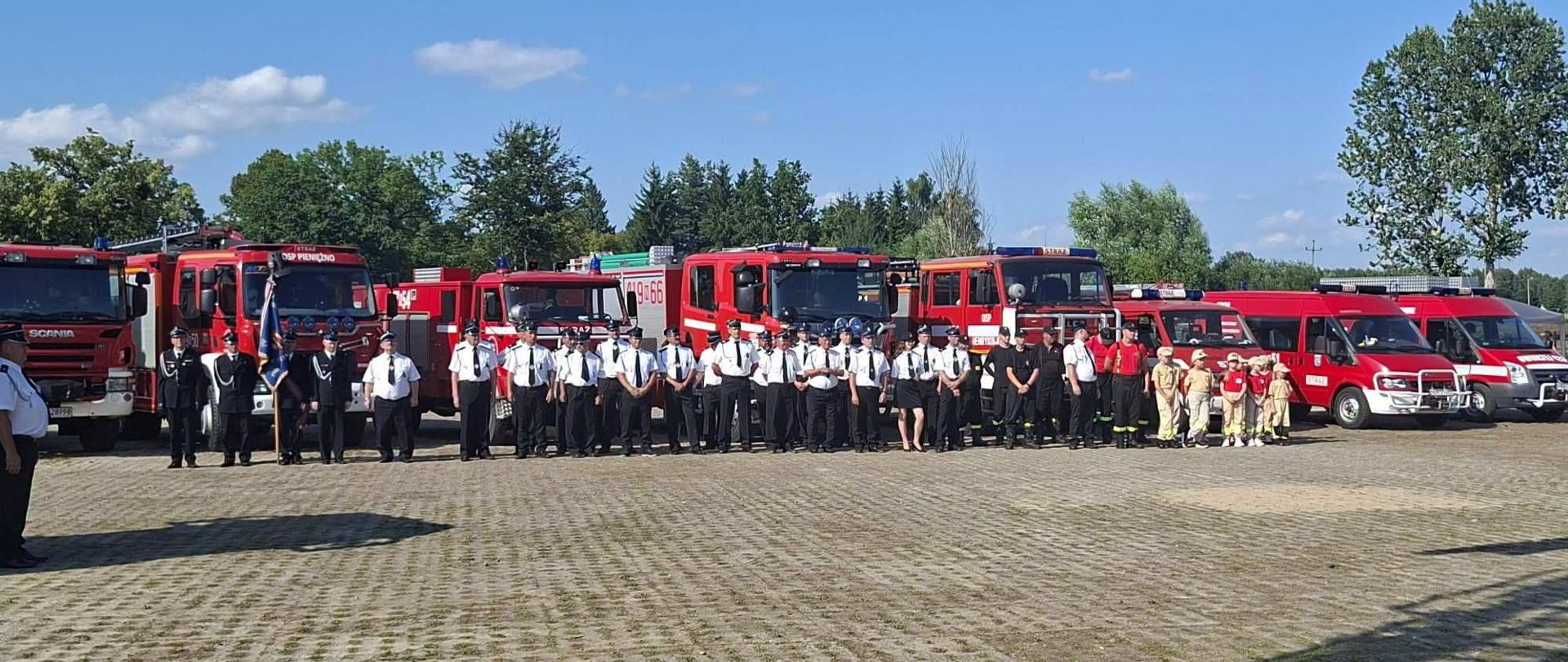 Na zdjęciu duża grupa strażaków ochotników w mundurach galowych, białych koszulach letnich. W tle za nimi czerwone samochody strażackie ustawione jeden obok drugiego.