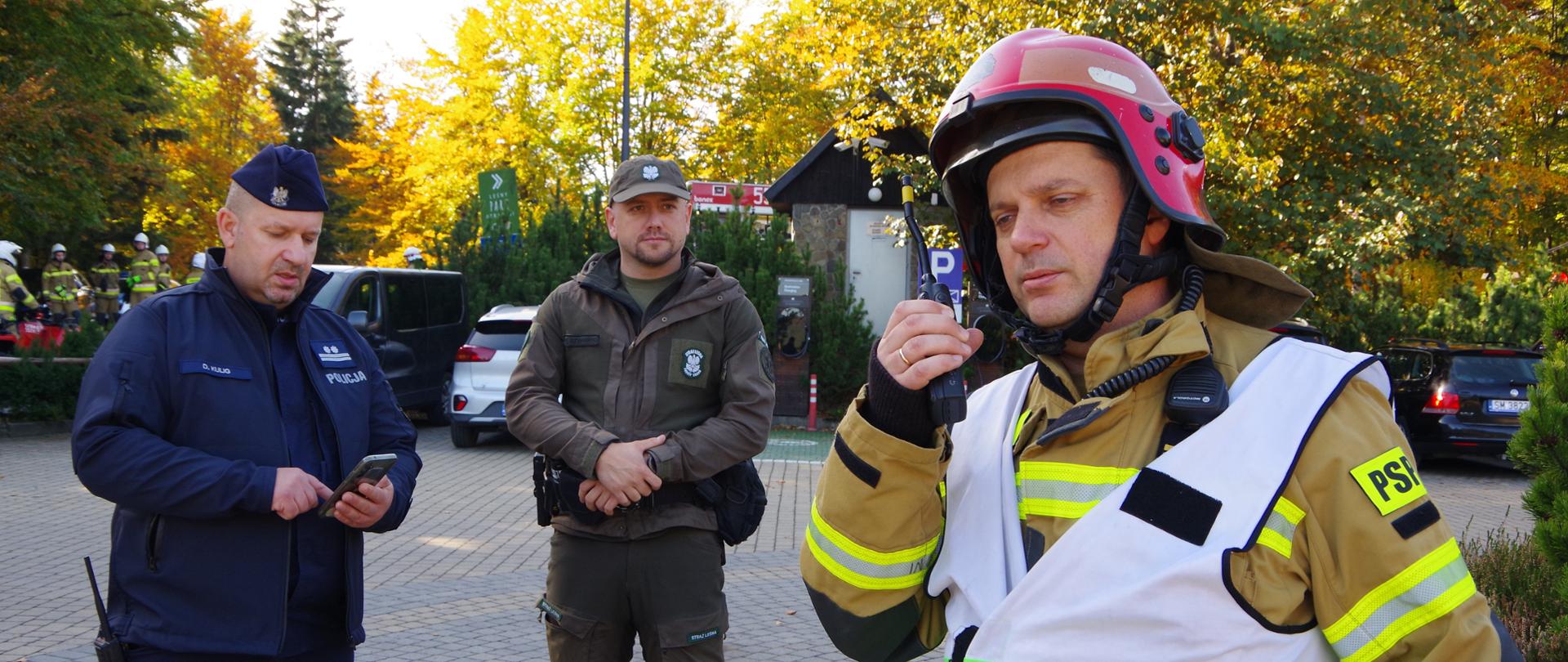 Widok z przodu. Od prawej kierujący działaniem ratowniczym na poziomie taktycznym strażak, nawiązujący korespondencję radiową za pomocą radiotelefonu. W środku funkcjonariusz straży leśnej, a po prawej policjant. W tle stanowisko ładowania pojazdów elektrycznych i otoczenie resortu hotelowego. 