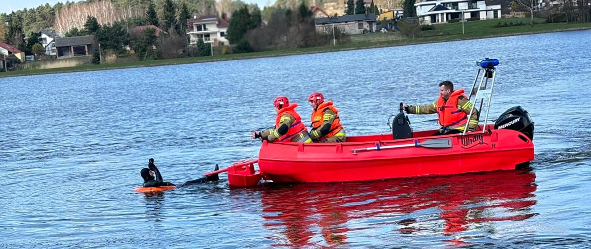 Na łódce ratowniczej znajduje się 3 strażaków ubranych w stroje strażackie i kamizelki ratunkowe. Obok łódki, w wodzie znajduje się człowiek symulujący topienie się w ramach ćwiczeń z ratownictwa wodnego.