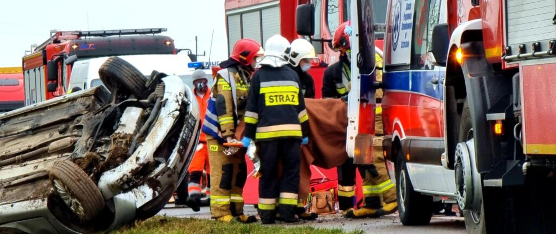 Strażacy pomagają w transporcie osoby poszkodowanej do karetki pogotowia. Widać auto leżące na dachu oraz pojazdy służb ratowniczych.