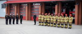 Zdjęcie wykonane na zewnątrz budynku na tle garaży. Przedstawia zbiórkę strażaków z okazji święta. Strażacy stoją w dwuszeregu w umundurowaniu specjalnym. 