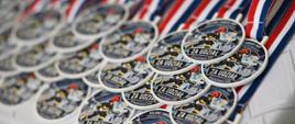 Zbliżenie na rząd pamiątkowych medali na biało-czerwono-niebieskich wstążkach. Na medalach widnieje ilustracja młodego strażaka w kasku oraz napis „ZA UDZIAŁ”.
