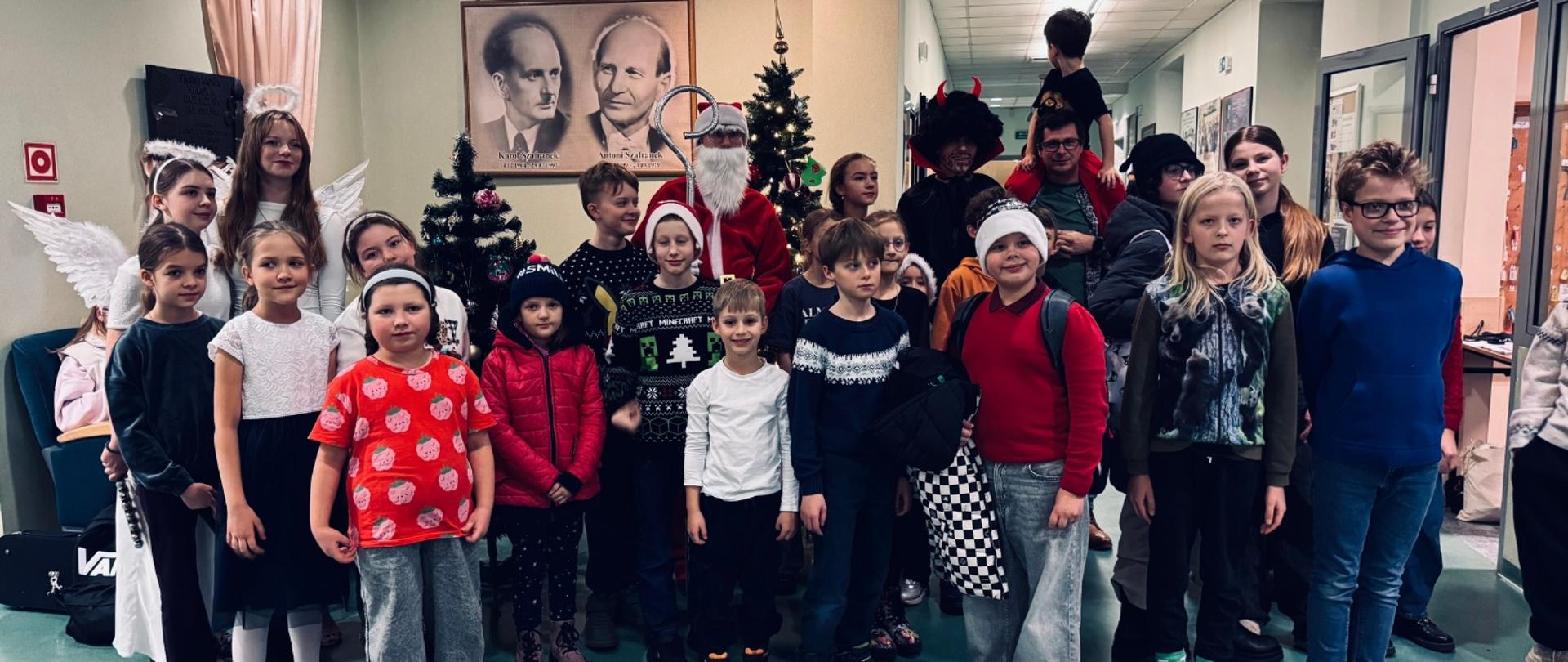 Na zdjęciu widać grupę dzieci i kilka osób dorosłych pozujących razem podczas strojenia choinki z Mikołajem. W tle stoją ozdobione choinki, a w centrum znajduje się Święty Mikołaj. Wśród uczestników są też osoby ubrane w stroje aniołów i diabłów. Dzieci mają na sobie zimowe ubrania, niektóre noszą czapki świąteczne. W tle widać korytarz szkolny oraz portrety patronów szkoły Karola i Antoniego Szafranków wiszące na ścianie. Cała scena wygląda radośnie i świątecznie.