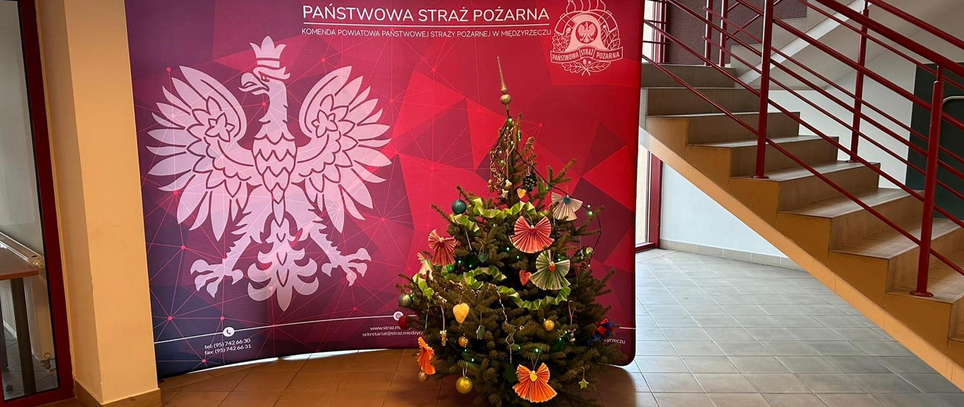 Zdjęcie przedstawia świąteczną choinkę w holu komendy powiatowej PSP w Międzyrzeczu na tle logo oraz godła państwowego