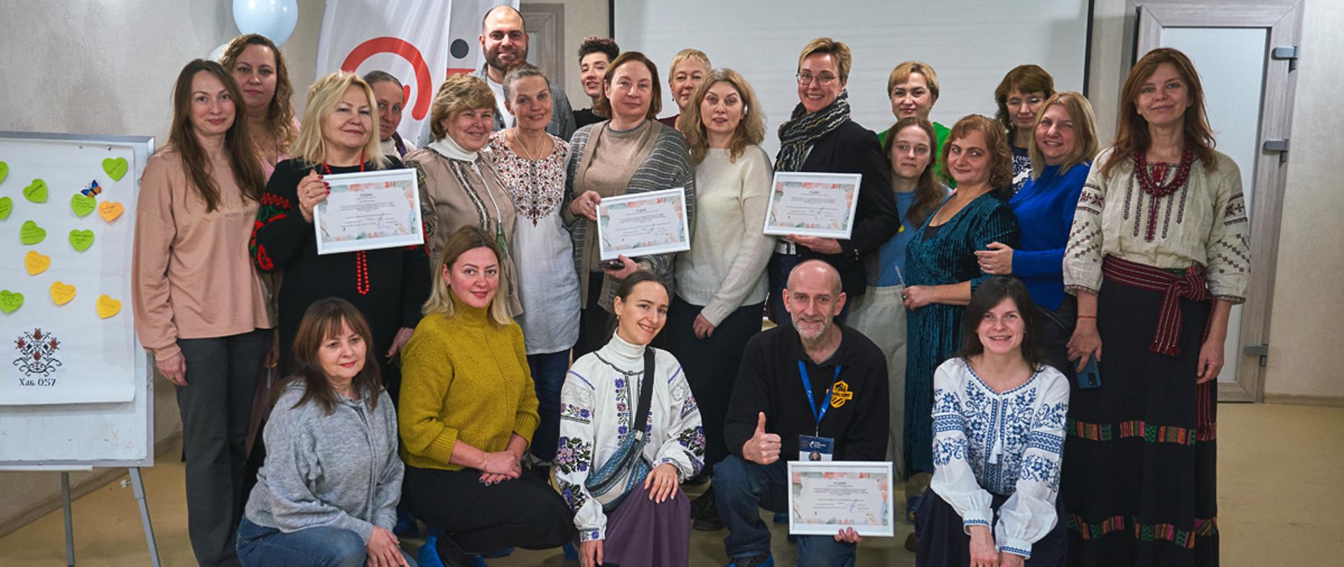 Grupa kobiet, część w ukraińskich strojach ludowych, prezentuje certyfikaty, w tle widać zarys logo Polskiej pomocy