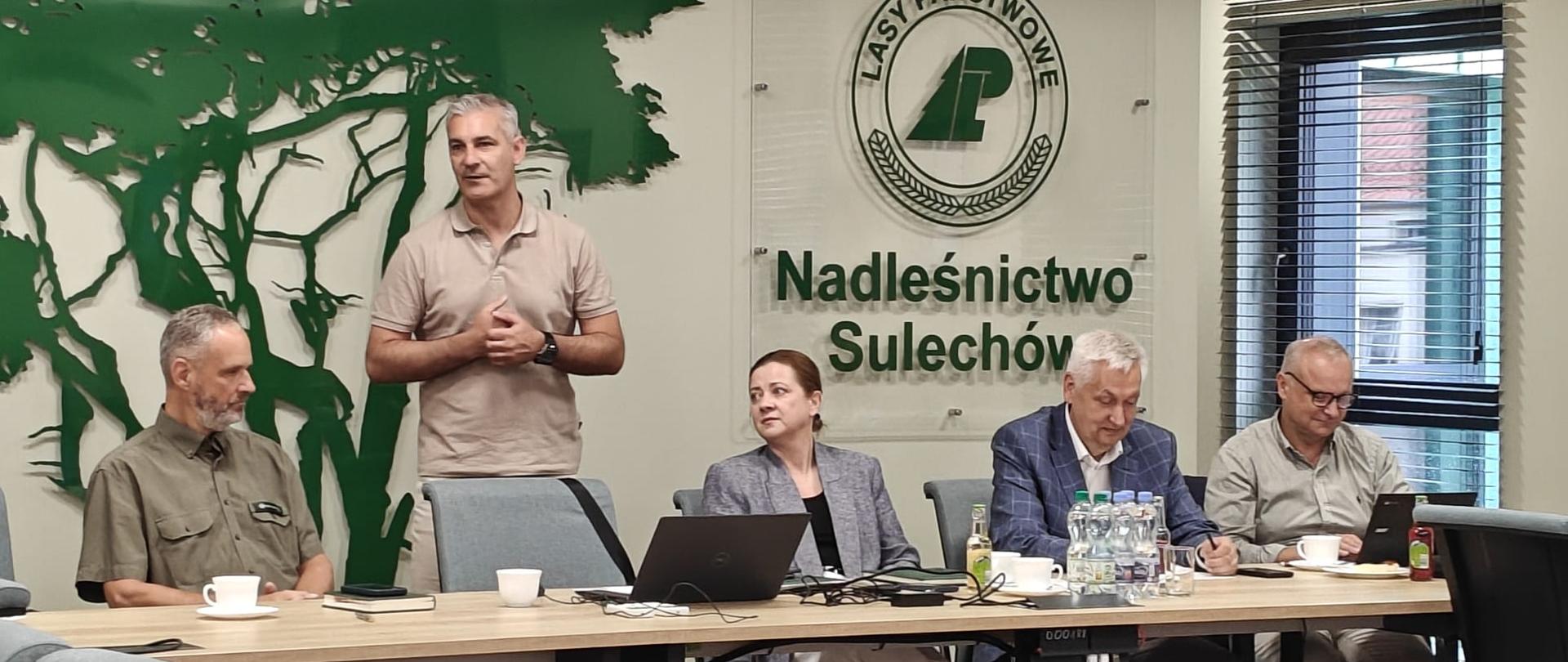 Na zdjęciu przy stole ustawionym w kształcie litery U siedzą przedstawiciele RDOŚ w Gorzowie Wlkp. Pani Agnieszka Nowak - Regionalny Dyrektor Ochrony Środowiska w Gorzowie Wielkopolskim po lewej stronie stojąc przemawia Pan Michał Bielewicz - Zastępca Regionalnego Dyrektora Ochrony Środowiska w Gorzowie Wielkopolskim. Po obu stronach siedzą członkowie RROP. Za plecami napis-baner Nadleśnictwo Sulechów a na stołach tablet , filiżanki i woda 