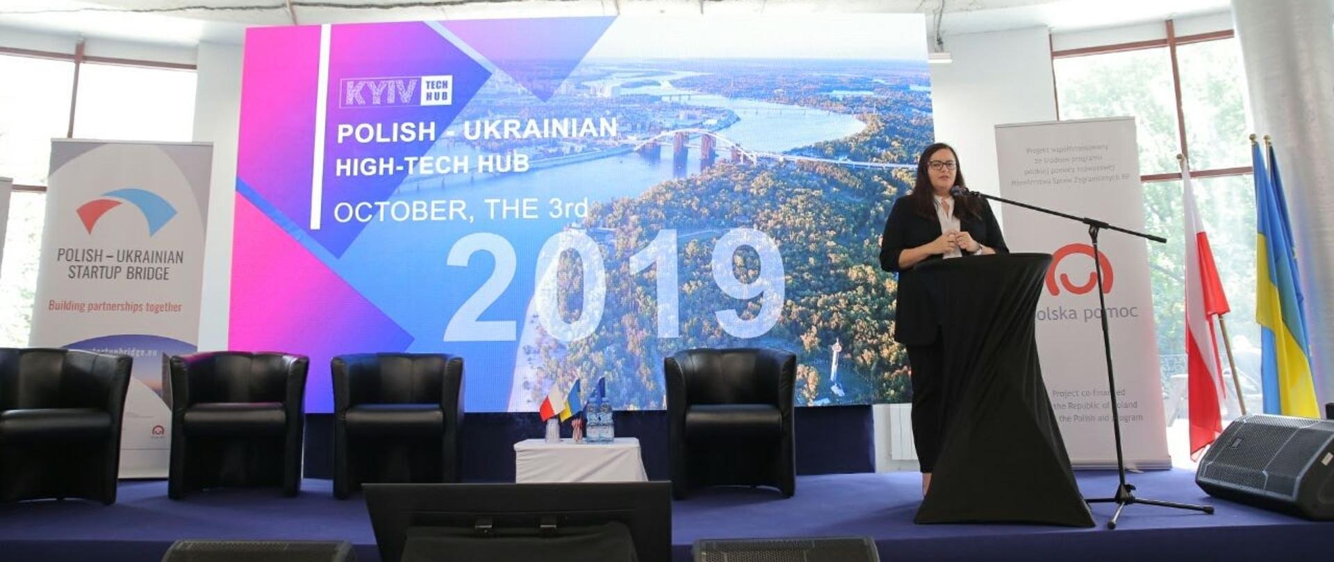 Wiceminister Małgorzata Jarosińska-Jedynak podczas otwarcia konferencji na scenie przy czarnej mównicy, z tyłu ekran z grafiką promocyjną wydarzenia z napisem: "Kyiv Tech Hub, Polish-Ukrainian High-Tech Hub, October the 3rd 2019", po lewej stronie roll-ip z logo i napisem Polish-Ukrainian Startup Bridge, po prawej obok sceny flagi Polski i Ukrainy