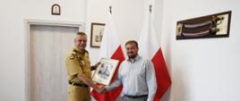 Funkcjonariusz Państwowej Straży Pożarnej trzyma dłoń oraz wręcza mężczyźnie obraz za nimi stoją dwie flagi Polski wiszą obraz oraz szabla na ścianie. 