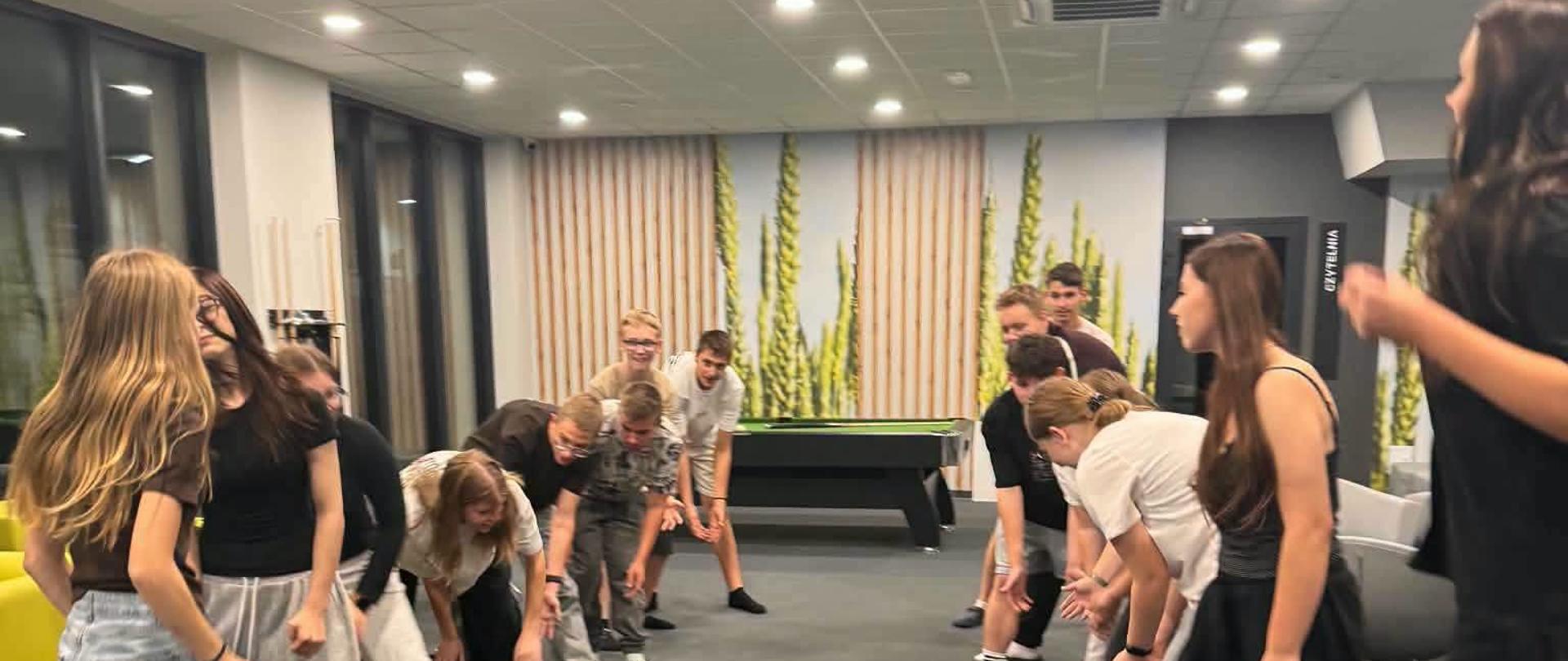 Grupa młodzieży w luźnych strojach uczestniczy w zajęciach ruchowych w przestronnej sali z bilardem w tle. Dwaj nastolatkowie w pierwszym planie patrzą na siebie, podczas gdy pozostali uczestnicy, ustawieni w dwóch rzędach, schylają się w rytmicznym ruchu. W tle widać dekoracyjny mural z bambusem i duże okna.