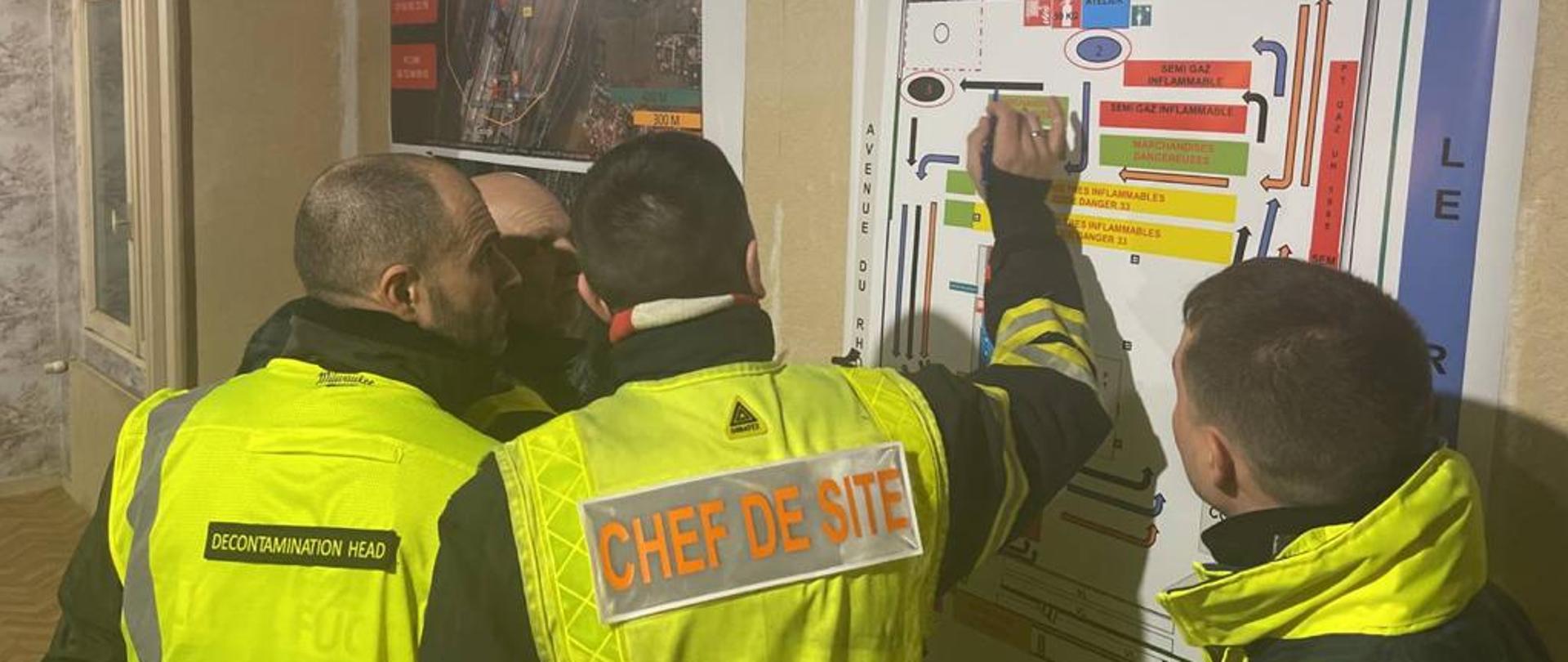 Czterech mężczyzn w odblaskowych kurtkach, jeden z napisem: chef de site stojących przy planie z informacjami zawieszonymi na ścianie. 