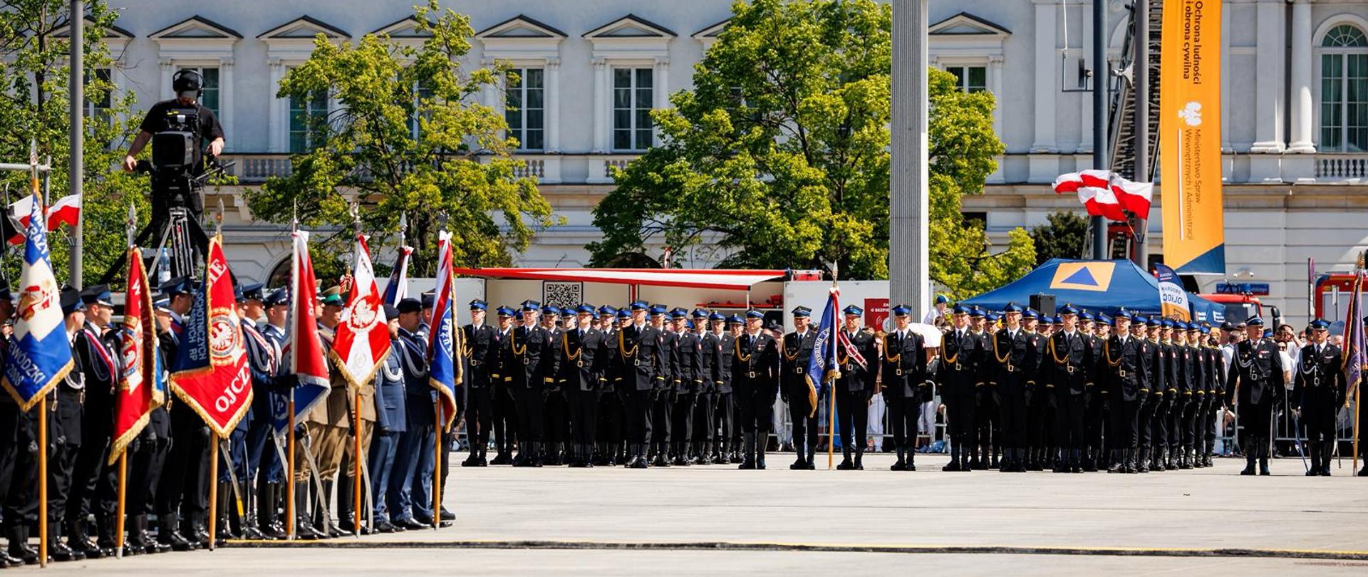 Panorama placu uroczystości z widocznymi pocztami sztandarowymi, zwartą formacją funkcjonariuszy oraz zabudową pałacową w tle.
