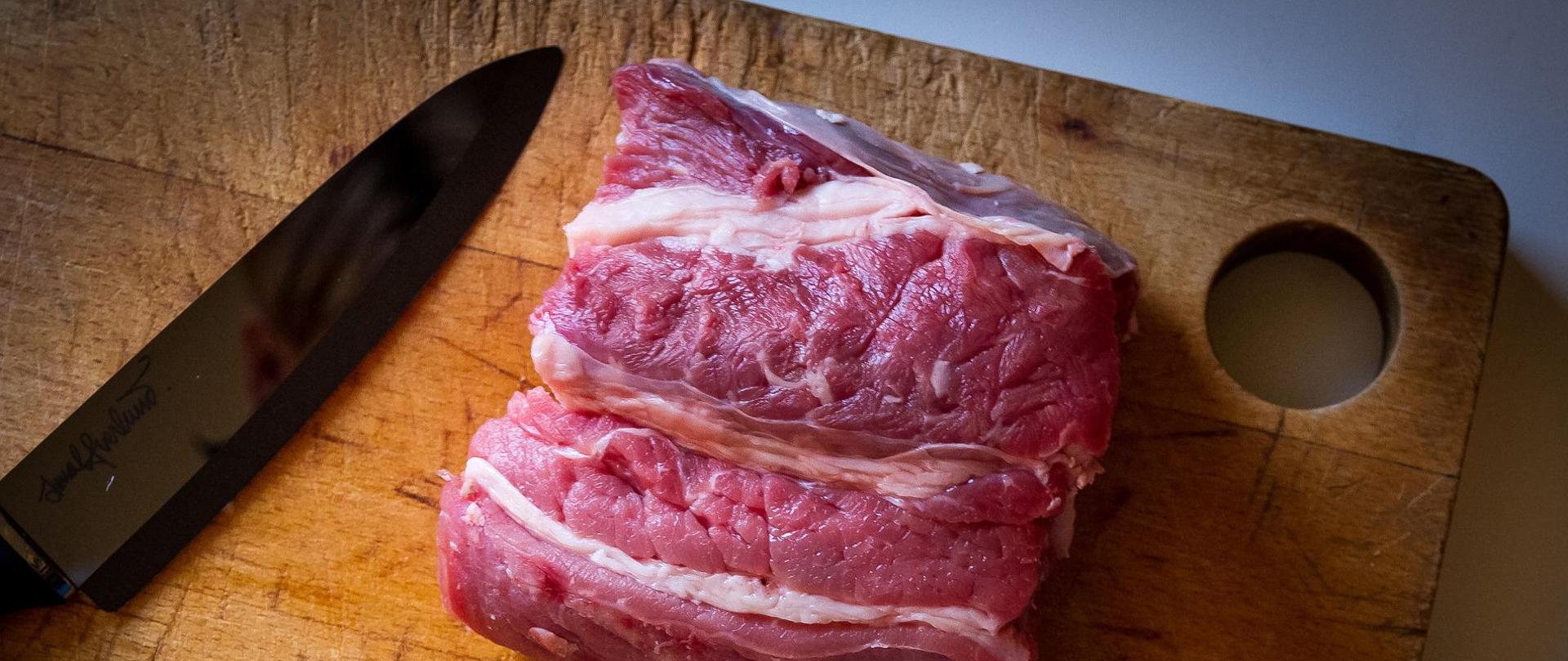 Na drewnianej desce znajduje się kawałek mięsa wołowego. Z lewej strony obok mięsa leży nóż.