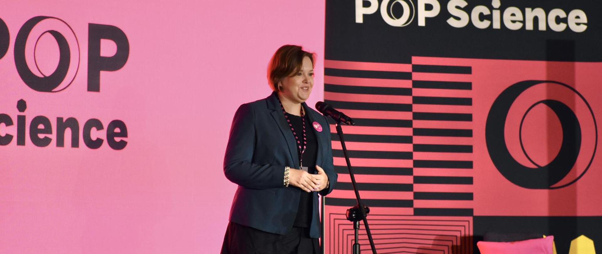Zdjęcie z boku pod kątem, wiceminister Zioło-Pużuk w ciemnoszarej marynarce stoi na tle różowo-fioletowej ściany z napisem POP Science i mówi do mikrofonu na stojaku.