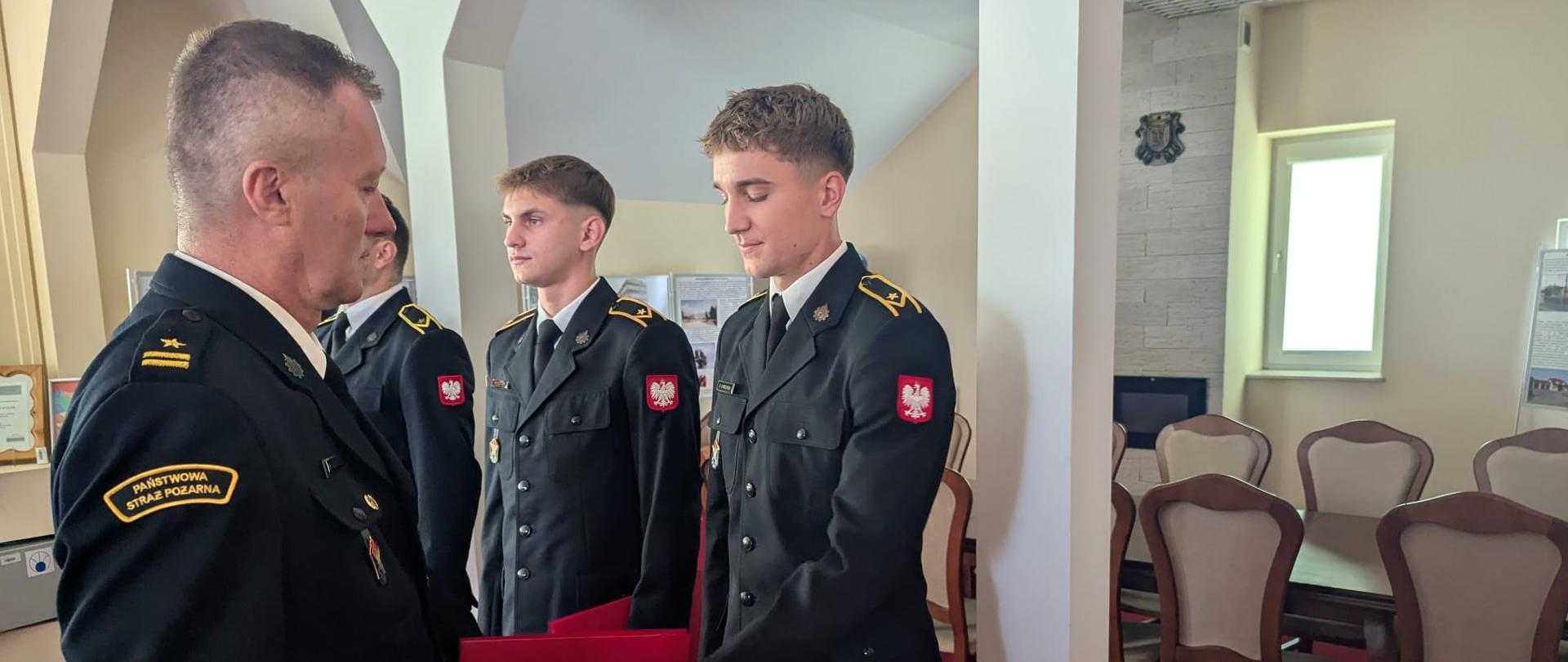 Z bliska, w jasnym pomieszczeniu z białymi kolumnami, starszy stopniem oficer straży pożarnej wręcza młodemu strażakowi w galowym mundurze czerwoną teczkę, podczas gdy w tle stoi kolejny młody strażak, a w głębi widać stół i krzesła w stylu klasycznym.