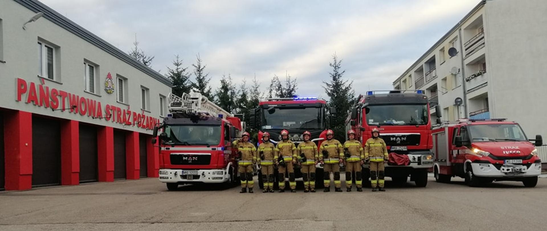Strażacy stoją w szeregu przed remizą. W samochodach widać włączone sygnały świetlne. 