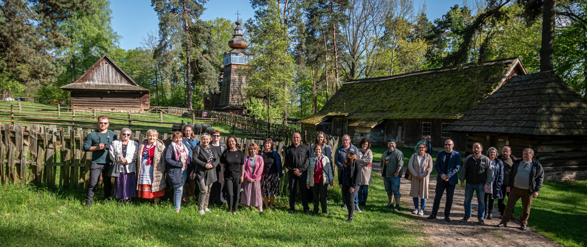 Zdjęcie przedstawia grupę ludzi, którzy stoją obok siebie i pozują do zdjęcie na tle starych drewnianych chat i wieży kościelnej oraz zieleni traw i drzew. 