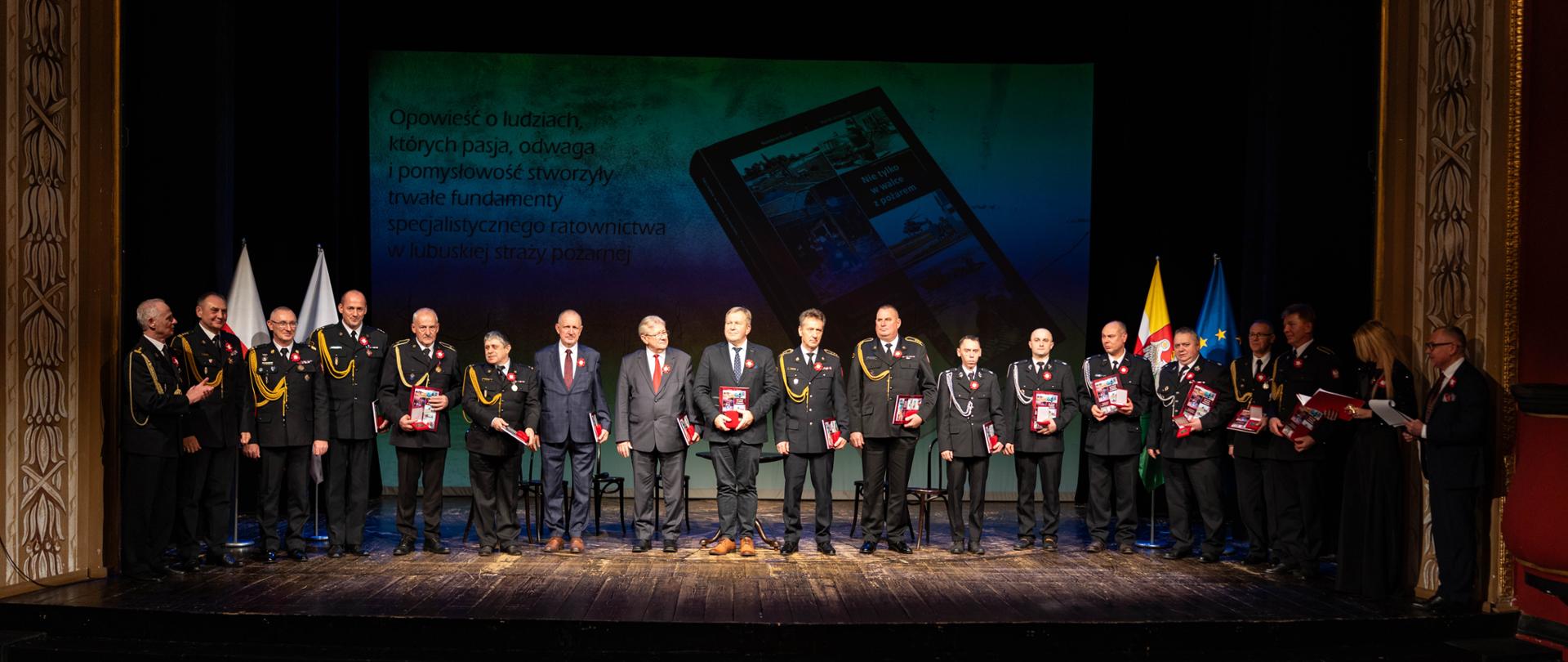 Grupa kilkunastu mężczyzn, zarówno w mundurach strażackich jak i w garniturach, stoi w rzędzie na scenie. Wszyscy trzymają w dłoniach egzemplarze książki. W tle duży ekran prezentujący okładkę publikacji z tytułem "Nie tylko w walce z pożarem – początki ratownictwa specjalistycznego w lubuskiej straży pożarnej". Po prawej stronie widać prowadzących uroczystość.