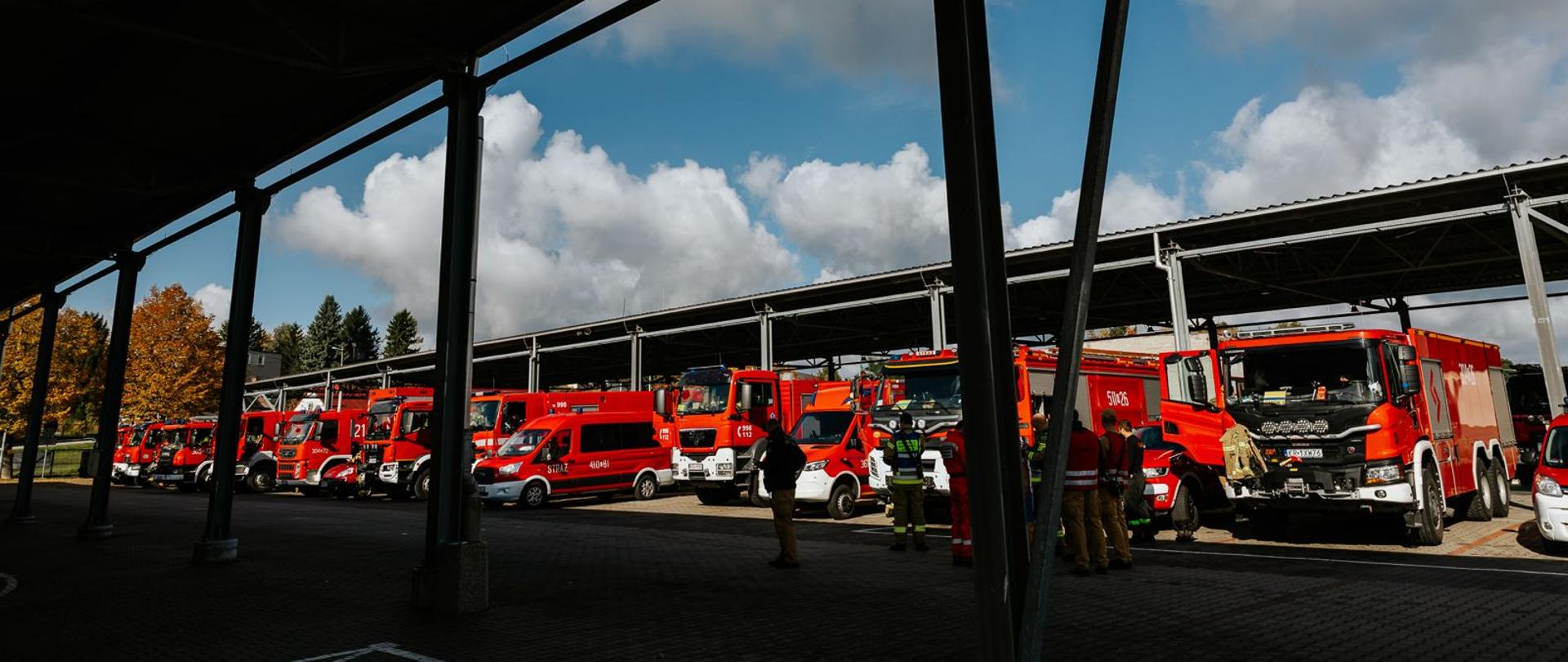 Szereg pojazdów ratowniczo-gaśniczych PSP zaparkowanych pod wiatą garażową. Na placu strażacy przygotowują się do rozpoczęcia działań lub ćwiczeń