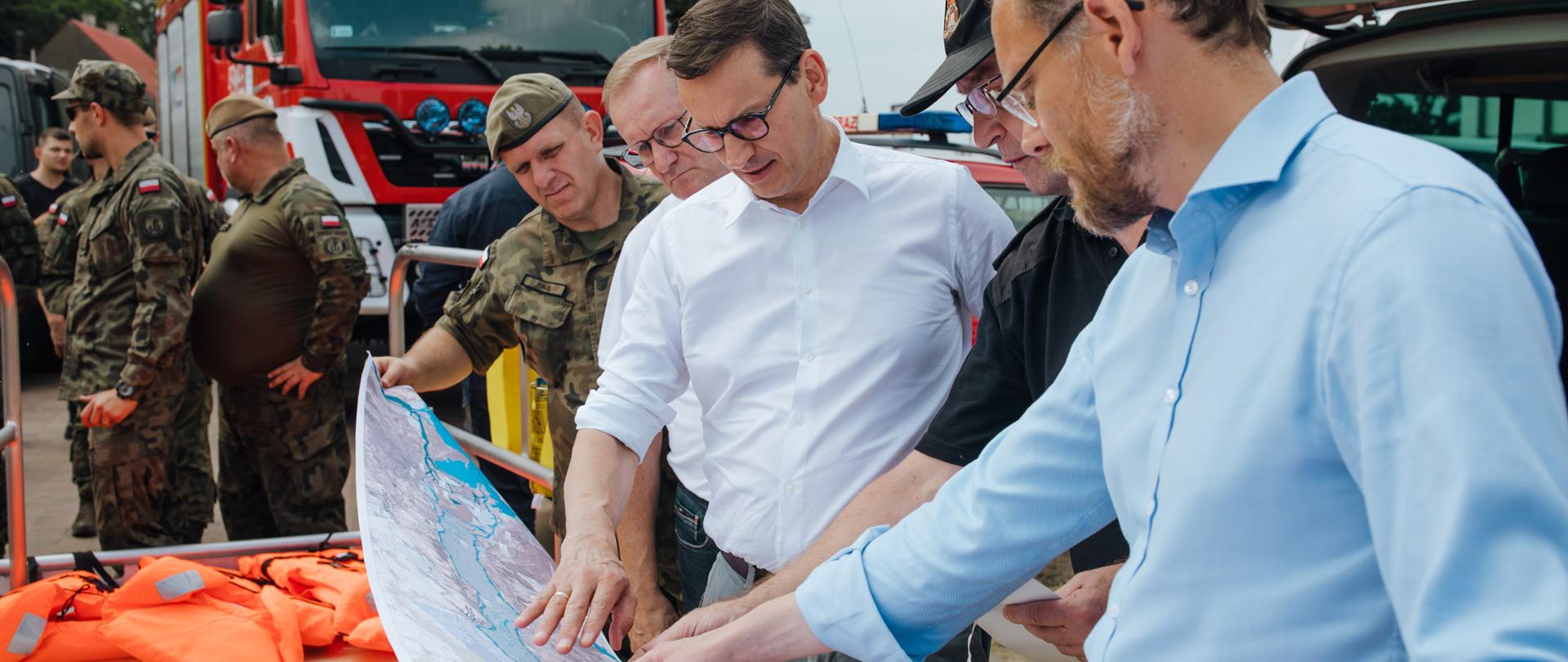 Premier Mateusz Morawiecki ogląda mapę, na drugim plaie wojsko oraz służby.