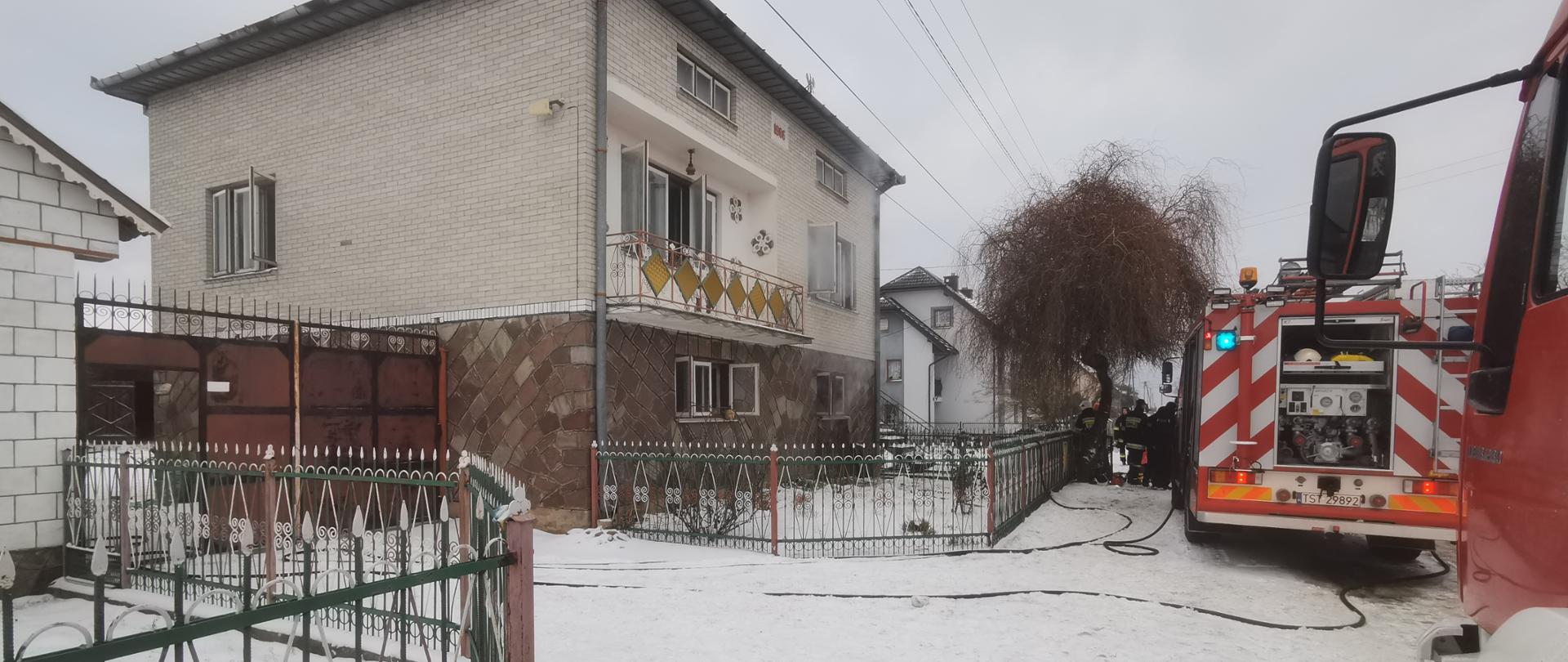 Budynek mieszkalny, murowany z białej cegły po pożarze, przed budynkiem z prawej strony samochody pożarnicze z rozwiniętą linią szybkiego natarcia i ratownicy PSP, OSP, pora dzienna